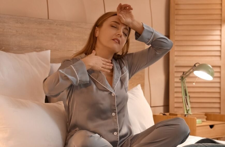 Mulher em pijama sentada na cama com expressão de desconforto, tocando o peito e a testa — uma cena comum entre os primeiros sinais da menopausa, como as ondas de calor noturnas.