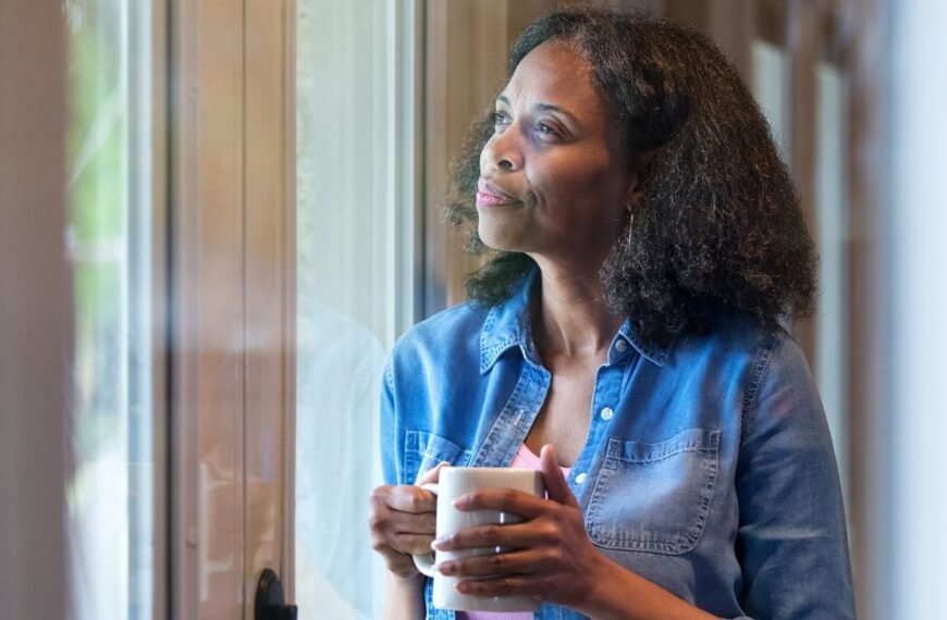 Mulher na faixa dos 50 anos olhando pela janela, em momento de reflexão sobre quanto tempo duram os sintomas da menopausa.
