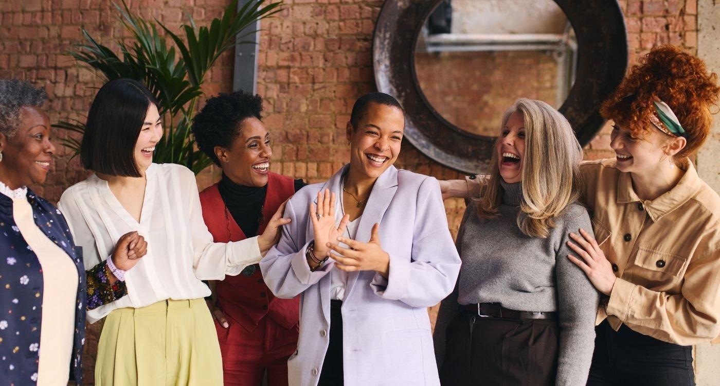 Grupo diverso de mulheres sorrindo e interagindo com alegria, representando acolhimento, diversidade e apoio mútuo durante a menopausa em diferentes culturas.