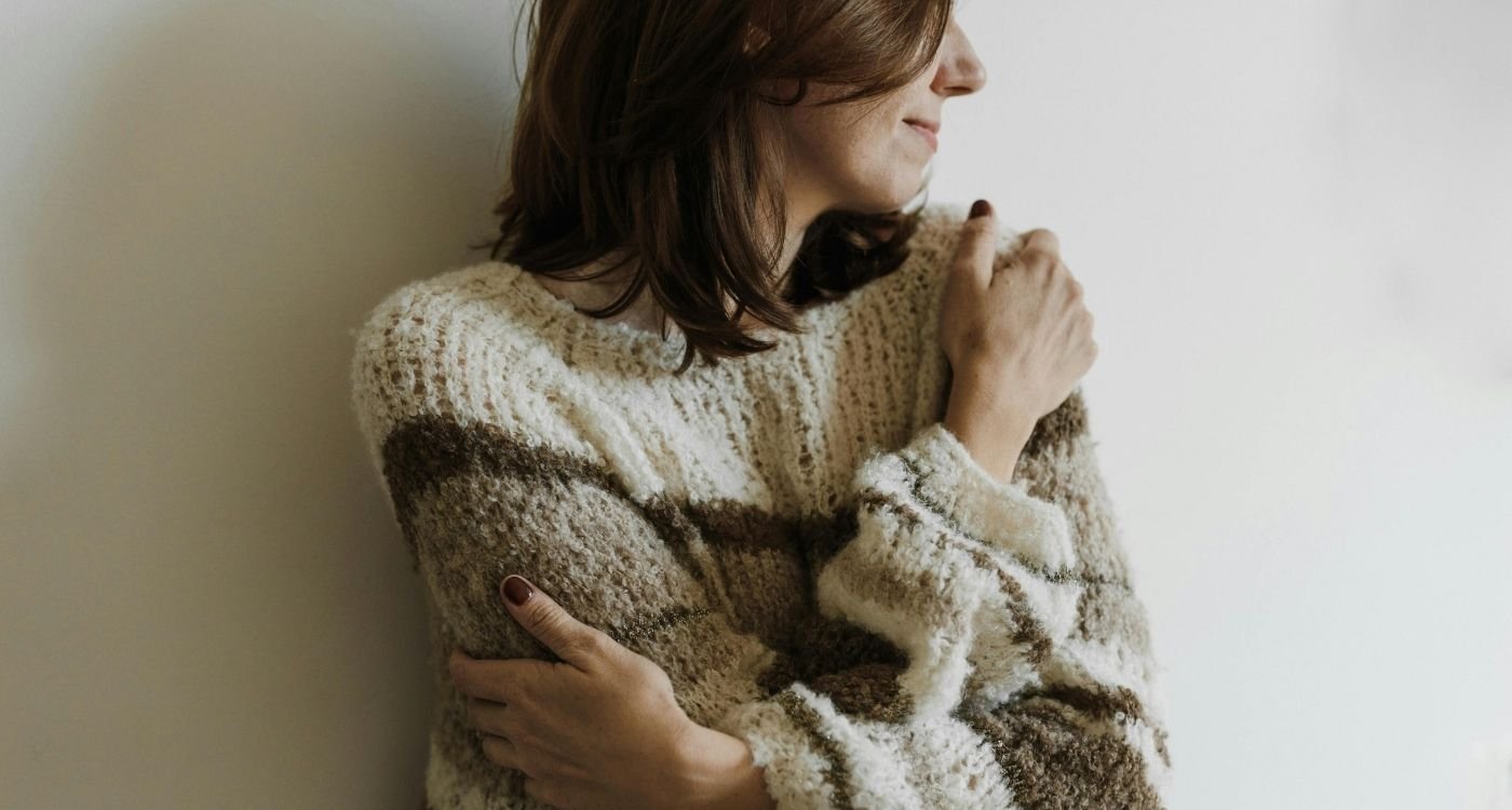 Mulher 50+ de perfil, abraçando‑se com carinho, suéter de tricô em tons neutros, luz lateral suave e fundo claro representando a autoestima na menopausa.