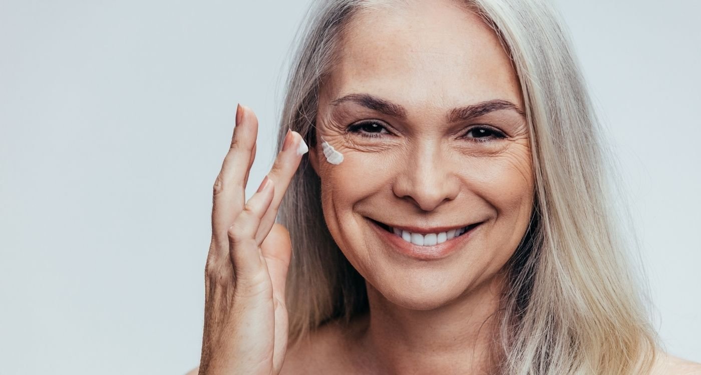 Mulher madura sorridente aplicando creme no rosto, representando cuidados com a pele e colágeno na menopausa.