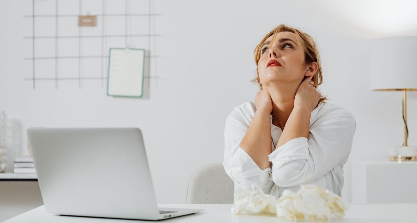 Menopausa no trabalho: mulher 50+ em escritório, mãos no pescoço e expressão de cansaço.