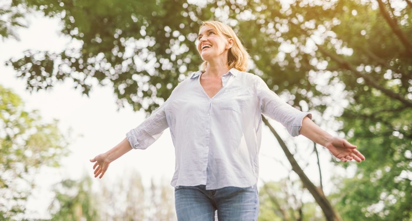 Mulher 50+ sorrindo e caminhando em parque, braços abertos, clima de primavera e bem-estar — viagem segura aos 50.