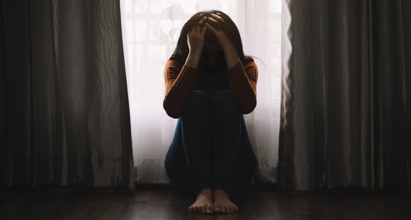 Mulher sentada no chão, próxima à janela, com as mãos na cabeça em sinal de sofrimento — representação da depressão na menopausa.