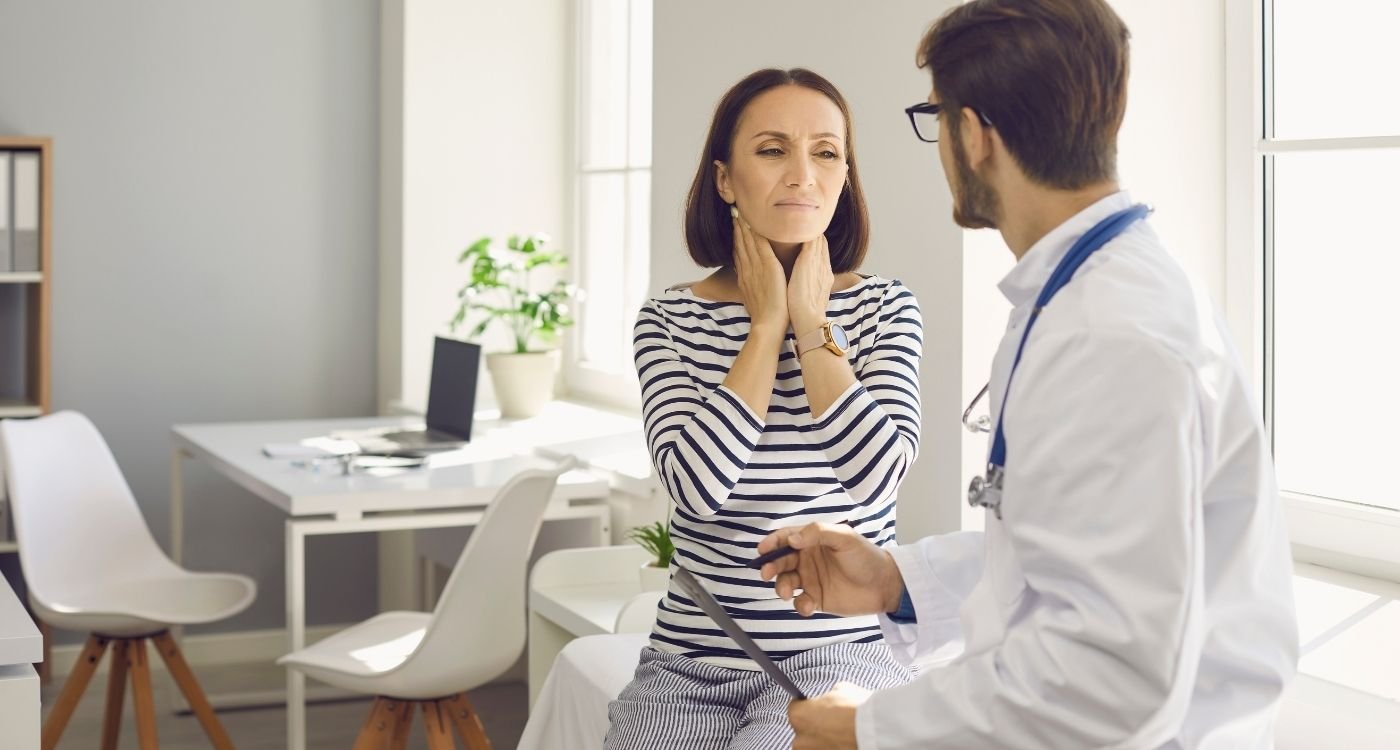 Mulher 50+ em consulta médica preventiva, tocando o pescoço, conversa com médico — menopausa silenciosa e autocuidado.