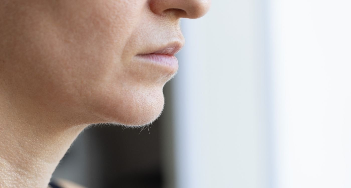 Mulher 40+ em close com pelos faciais na menopausa no queixo, luz natural e fundo claro.