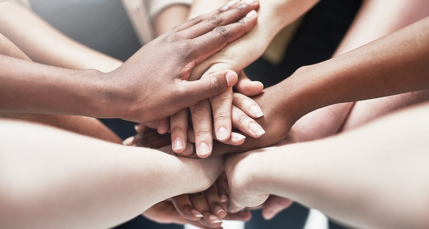 Mãos de mulheres 40+ de tons de pele diversos sobrepostas em gesto de união, simbolizando a rede de apoio na perimenopausa.