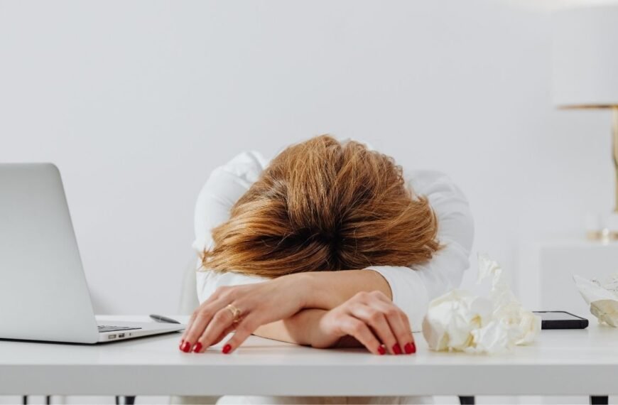 Mulher de meia-idade debruçada sobre a mesa do escritório, ao lado de um notebook, com postura exausta, representando o cansaço na menopausa no dia a dia de trabalho.