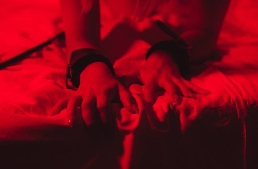 Viagra feminino: close-up de mãos femininas sobre a cama, com algemas nos pulsos, em iluminação vermelha, sugerindo sensualidade e fantasia de forma discreta.