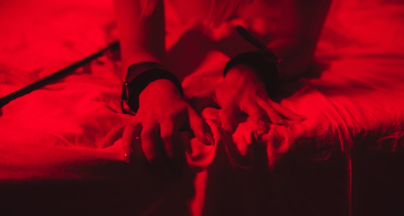 Viagra feminino: close-up de mãos femininas sobre a cama, com algemas nos pulsos, em iluminação vermelha, sugerindo sensualidade e fantasia de forma discreta.