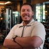 Eduardo Pinheiro, personal trainer para mulheres 40+, sorrindo de braços cruzados em uma academia, com foco em fortalecimento muscular, saúde e qualidade de vida.